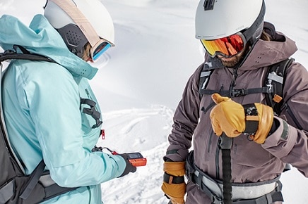 kobieta i mężczyzna ubrani w kaski i gogle narciarskie sprawdzają detektor lawinowy