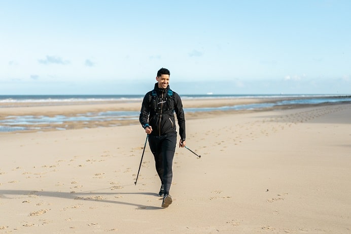 mężczyzna idący w stroju sportowym po plaży z kijkami do nordic walking w rękach